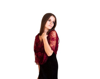 May include: A woman wearing a burgundy lace shawl over a black dress. The shawl has a delicate floral pattern and a flowing design.