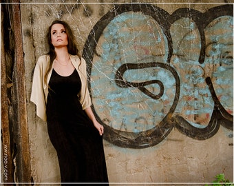 May include: A woman wearing a black dress and a beige shawl stands in front of a graffiti-covered wall. The wall is textured and has a blue, black, and white color scheme.
