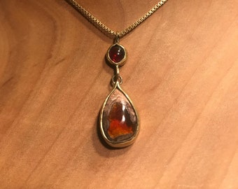 May include: A gold-colored necklace with a teardrop-shaped pendant and a smaller, round stone above it. The pendant has a fiery orange and red center, and the chain is a delicate box chain.