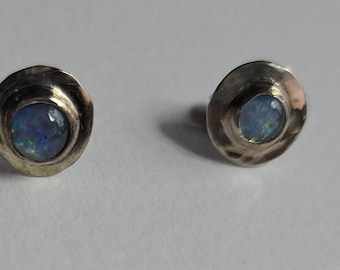 May include: A pair of stud earrings with a silver-colored setting. Each earring features a round, iridescent blue opal stone. The earrings have a textured, wavy design around the stone.
