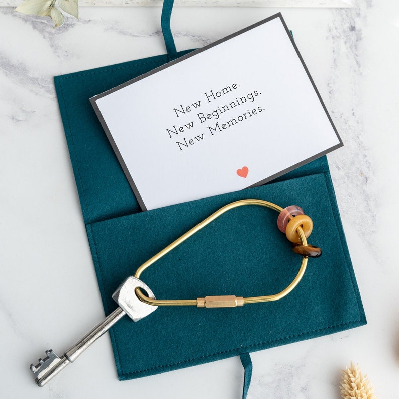 May include: Teal felt key holder with brass key ring and multicolored beads.  A gold key is attached.  An accompanying card reads: 'New Home. New Beginnings. New Memories.'  The overall theme is a new home gift.