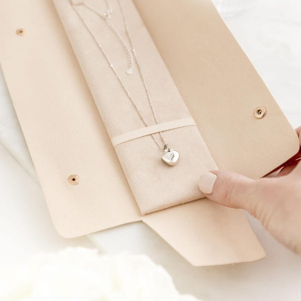 May include: A silver necklace with a heart-shaped pendant is displayed inside a light beige leather jewelry case. The case is open, revealing the necklace nestled in a soft, cream-colored interior. The pendant has a small initial engraved on it.