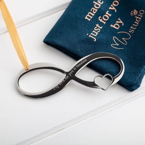 Peut inclure: Symbole d'infini en argent avec une découpe en forme de cœur et l'inscription "My love for you is timeless". Le symbole est sur une surface blanche et est livré avec une pochette en velours bleu avec le texte "made just for you by M studio".