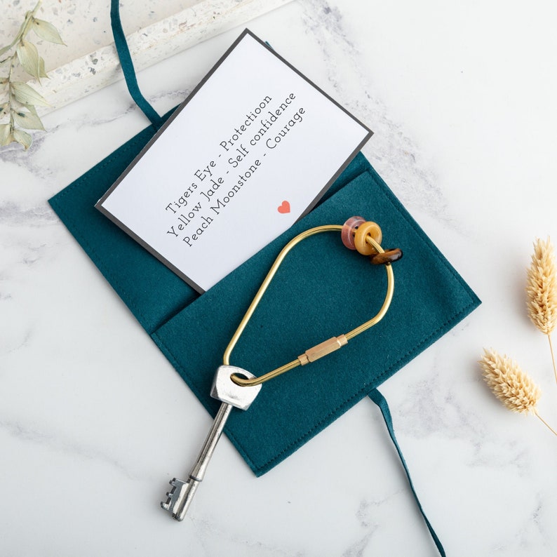 May include: Teal felt pouch containing a brass key ring with assorted gemstone beads and a card describing the stones: Tigers Eye - Protection, Yellow Jade - Self Confidence, Peach Moonstone - Courage. The key ring is attached to a silver key.