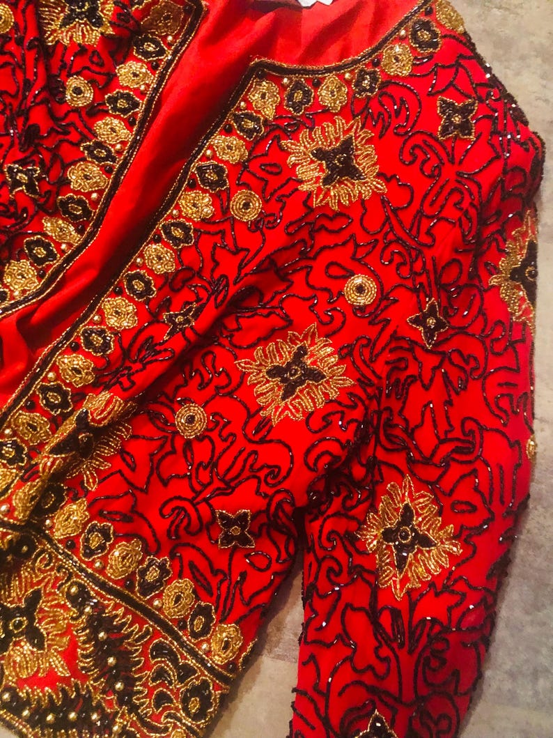May include: A red jacket with intricate gold and black beadwork. The jacket features floral and scroll patterns, with a V-neckline and long sleeves. The embellishments create a textured, ornate design.