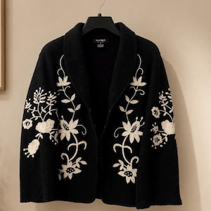 May include: A black open-front cardigan featuring a shawl collar and long sleeves. The garment is decorated with white floral embroidery that extends from the hem up the front panels and across the sleeves. A black hanger is attached at the top.
