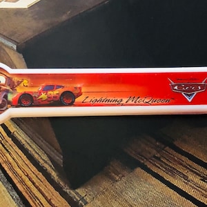 Puede incluir: Un utensilio de acero inoxidable con un mango rojo y blanco que presenta personajes y texto de la película Cars de Disney/Pixar. El mango tiene imágenes de Mate y Rayo McQueen, con el texto "Lightning McQueen".