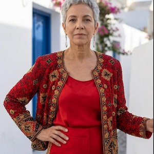 May include: A woman in a red outfit, including a top and pants, stands outdoors. She wears a red jacket with gold embroidery. The background features white buildings, a blue door, and pink flowers.