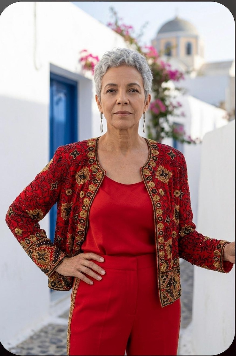 May include: A woman in a red outfit, including a top and pants, stands outdoors. She wears a red jacket with gold embroidery. The background features white buildings, a blue door, and pink flowers.