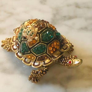 May include: A decorative turtle figurine, featuring a gold-coloured metal frame. The shell is adorned with green, orange, and brown gemstones. The turtle's head and feet are also gold, with red gemstones for eyes.