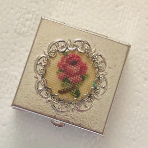 Peut inclure: Un boîtier compact carré en métal argenté avec un insert circulaire central. L'insert présente un motif floral en point de croix représentant une rose rouge avec des feuilles vertes sur fond crème, encadré par une bordure en métal argenté ajourée.