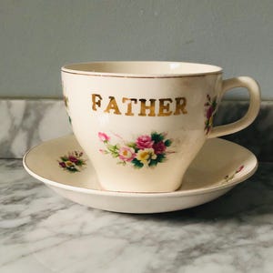 May include: Cream-colored teacup and saucer set. The teacup features the gold word "FATHER" and floral accents in pink, yellow, and green. The saucer has matching floral details. The set is likely vintage.