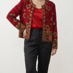 May include: A red beaded jacket with gold and black floral embroidery. The jacket is worn over a red top and paired with black pants and loafers. The outfit is a stylish ensemble.
