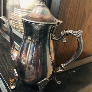 May include: A silver antique teapot with a lid and ornate handle. The teapot has a curved spout and sits on three decorative feet. The surface reflects the surrounding environment.