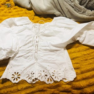 May include: A white blouse with a round neckline, long, puffy sleeves, and a lace-up front. The bottom hem features a floral eyelet design. The blouse is made of a lightweight fabric and is laying on a yellow surface.