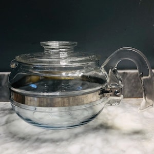 May include: A clear glass teapot with a stainless steel band and handle. The teapot has a round body and a lid. The handle is arched and attached to the side of the pot. The words "STAINLESS" are visible on the band.
