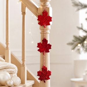 Peut inclure: Trois décorations rouges en forme de feuilles d'érable suspendues à un poteau d'escalier en bois. Les feuilles ont une surface texturée et sont attachées à une ficelle rouge et blanche. Une guirlande de petites lumières blanches est enroulée autour du poteau.