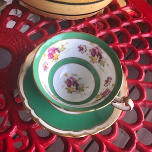 May include: A vintage teacup and saucer set. The teacup has a white interior with floral designs in purple, yellow, and pink. The exterior is green with a gold trim, matching the saucer. The set is placed on a red, ornate table.