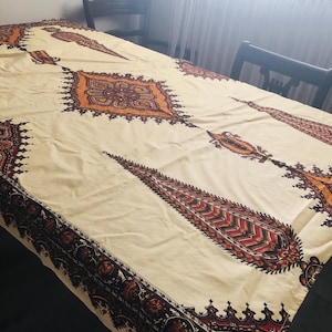 May include: Cream-coloured tablecloth with an intricate pattern in shades of brown, red, and orange. The design features geometric shapes and paisley-like motifs. The tablecloth is draped over a wooden table.