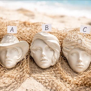 Peut inclure: Trois sculptures de visages en plâtre blanc cassé, chacune avec des coiffures et des couvre-chefs uniques, reposent sur un filet de pêche beige. Les sculptures sont étiquetées A, B et C. L'arrière-plan montre une scène de plage avec du sable et un aperçu de l'océan.