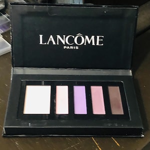 Puede incluir: Una paleta de sombras de ojos negra con el logo Lancôme Paris en blanco. La paleta contiene cinco tonos: un rosa claro, un rosa medio, un lavanda, un rosa más oscuro y un marrón.