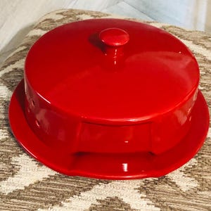 Peut inclure: Un plat à fromage en céramique rouge vif, avec un couvercle et un petit bouton sur le dessus. Le plat a une base ronde et un bord légèrement surélevé. Le couvercle s'adapte parfaitement, créant un récipient fermé.