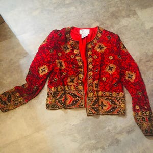 May include: A red jacket with intricate gold and black embroidery. The jacket features a round neckline and long sleeves. The embroidery includes floral and geometric patterns. The jacket is open in the front, revealing a red lining.