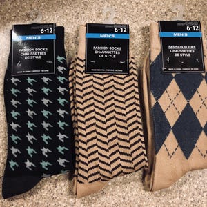 Puede incluir: Tres pares de calcetines de moda con etiquetas. Un par es negro con un patrón turquesa y blanco, otro es beige con un patrón en zigzag negro, y el último es beige con rombos azul marino. Cada par tiene una etiqueta que dice "MEN'S FASHION SOCKS CHAUSSETTES DE STYLE".