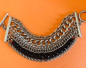 May include: Silver-tone chain bracelet with multiple strands. The bracelet features various chain styles, including rhinestone accents and a row of black beads. The clasp is a T-bar closure. The background is orange.