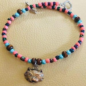 Peut inclure: Un bracelet de perles avec un charm en forme de chat. Le bracelet est composé de perles roses, bleues, marron et vert foncé. Le charm en forme de chat est de couleur bronze. Fermoir à bascule argenté.