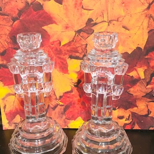 May include: A pair of clear glass candle holders with a geometric design. The candle holders have a wide, multi-faceted base and a detailed upper section to hold candles. The background features autumn leaves.