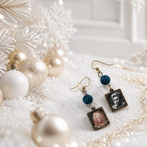 May include: A pair of dangle earrings featuring rectangular bronze-toned frames containing black and white portraits of a person. Each earring includes a dark blue textured spherical bead suspended from a gold-toned hook wire finding.