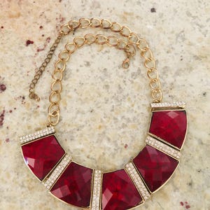 Op de afbeelding: Een opvallende ketting met grote, facetgeslepen, robijnrode stenen gezet in een goudkleurig metalen frame. De ketting heeft een gouden ketting met verstelbare lengte en is versierd met heldere strass-steentjes.