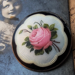 May include: A vintage silver and black brooch with a hand-painted pink rose and green leaves on a white background.