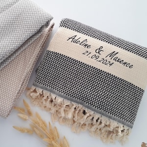 Puede incluir: Toallas grises y beige plegadas con estampado, junto a una toalla con estampado en blanco y negro con una franja color crema. La toalla en blanco y negro tiene el texto bordado "Adeline & Maxence 21.09.2024" y un borde con flecos.