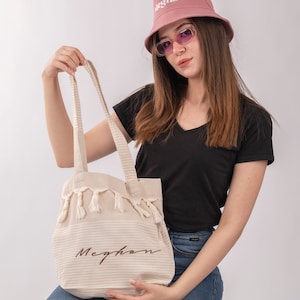 May include: A white tote bag with brown striped fabric and tassels. The bag has the name "Meghan" embroidered on it in brown thread. A person is holding the bag and wearing a pink bucket hat with the word "Meghan" embroidered on it in white thread.