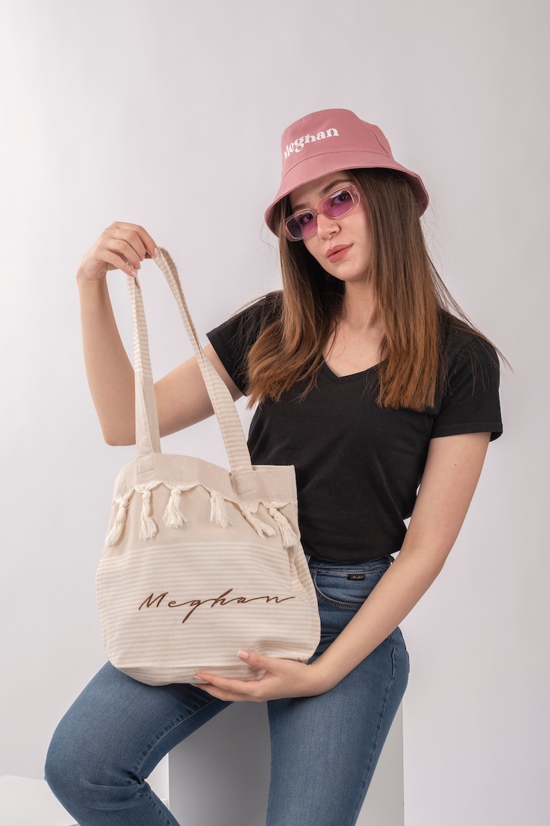 May include: A white tote bag with brown striped fabric and tassels. The bag has the name "Meghan" embroidered on it in brown thread. A person is holding the bag and wearing a pink bucket hat with the word "Meghan" embroidered on it in white thread.