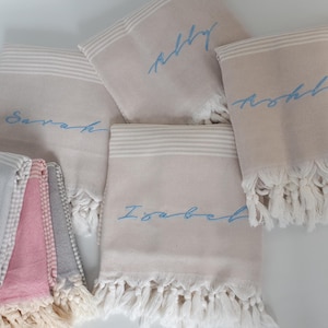 May include: Five personalized towels with fringe edges. The towels are beige with white stripes and have blue embroidered names: "Abby", "Noah", "Sarah", "Isabel", and "Ashley".