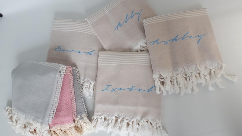 May include: Five personalized towels with fringe edges. The towels are beige with white stripes and have blue embroidered names: "Abby", "Noah", "Sarah", "Isabel", and "Ashley".