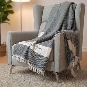 Può includere: Una poltrona grigia con una coperta a fantasia bianca e nera. La coperta ha strisce bianche, frange e il testo "& Maxence 09.09.2024". La coperta è drappeggiata sulla poltrona.