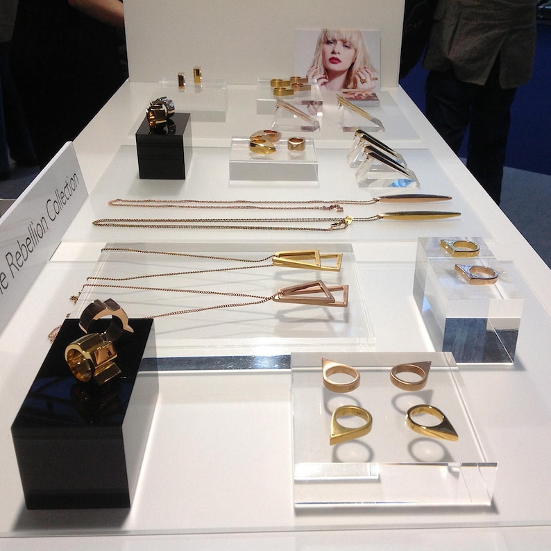 May include: A display of gold and rose gold jewelry on a white surface. The jewelry includes necklaces, rings, and earrings. The jewelry is arranged in a geometric pattern. The text "The Rebellion Collection" is visible in the lower left corner of the image.