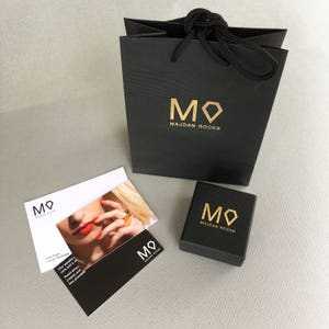 May include: A black gift bag with gold lettering that reads "MO MAJDAN ROCKS". A smaller black box with gold lettering that reads "MO MAJDAN ROCKS" is in front of the bag. A white card with gold lettering that reads "MO MAJDAN ROCKS" is in the background.