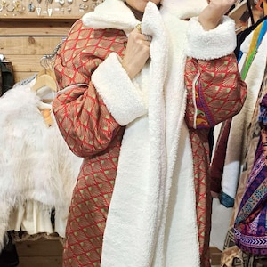 Peut inclure: Long manteau à motifs rouges et dorés, orné d'une bordure en fausse fourrure blanche sur le col, les poignets et le devant. Le manteau a un look douillet et surdimensionné, parfait pour superposer les couches. Les manches ont une forme arrondie unique.