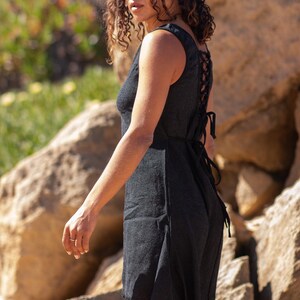 Black sleeveless hi-low ethnic dress in handwoven rustic cotton with round neckline, intentional distressed frayed detail at chest and dramatic asymmetric skirt short at front and long at back with raw frayed hem, worn against rocky coastal background - Raja Pura artisan boho dress - full body side view