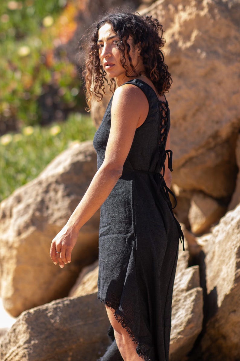 Black sleeveless hi-low ethnic dress in handwoven rustic cotton with round neckline, intentional distressed frayed detail at chest and dramatic asymmetric skirt short at front and long at back with raw frayed hem, worn against rocky coastal background - Raja Pura artisan boho dress - full body side view