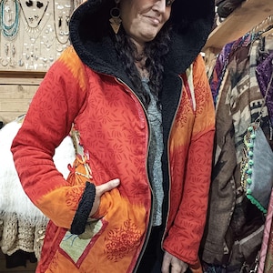Peut inclure: Un manteau à capuche réversible rouge et orange avec une doublure en polaire noire. Le manteau présente un motif floral et une fermeture éclair. Le modèle porte le manteau dans un magasin, avec des bijoux et d'autres vêtements en arrière-plan.