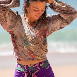 Peut inclure: Un chemisier cache-cœur à manches longues sur fond beige avec un motif floral dans les tons rouge, vert et marron, associé à une jupe paréo violette vive ornée d'un motif de feuilles tropicales et de fleurs vertes et roses.