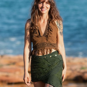 May include: A brown halter top with fringe and a green wrap skirt. The skirt has a layered design with decorative buttons. The outfit is worn outdoors with a blue sea in the background.