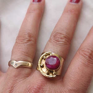 Könnte beinhalten: Zwei goldene Ringe werden auf einer Hand gezeigt. Einer ist ein schlichtes Band mit einem kleinen Diamanten. Der andere ist ein breiter, strukturierter Ring mit einem großen, ovalen, roten Edelstein und goldenen Details. Der Text "size 14" ist sichtbar.