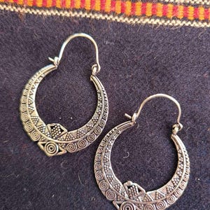 Peut inclure: Une paire de créoles argentées présentant un motif en relief complexe composé de spirales, de points et de triangles géométriques. Les boucles d'oreilles ont une forme incurvée en croissant et sont dotées d'une fermeture à charnière.